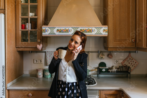 Smiling woman holding a coffee cup while talking on the phone in the kitchen at home.