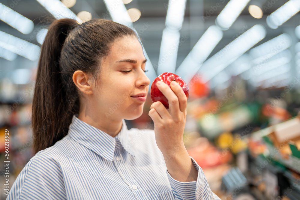 Young Caucasian woman enjoying the smell of a fresh apple. Woman smelling a red apple with the eyes closed while standing in a grocery store. Young Caucasian woman enjoying the smell of a fresh apple. Woman smelling a red apple with the eyes closed while standing in a grocery store.