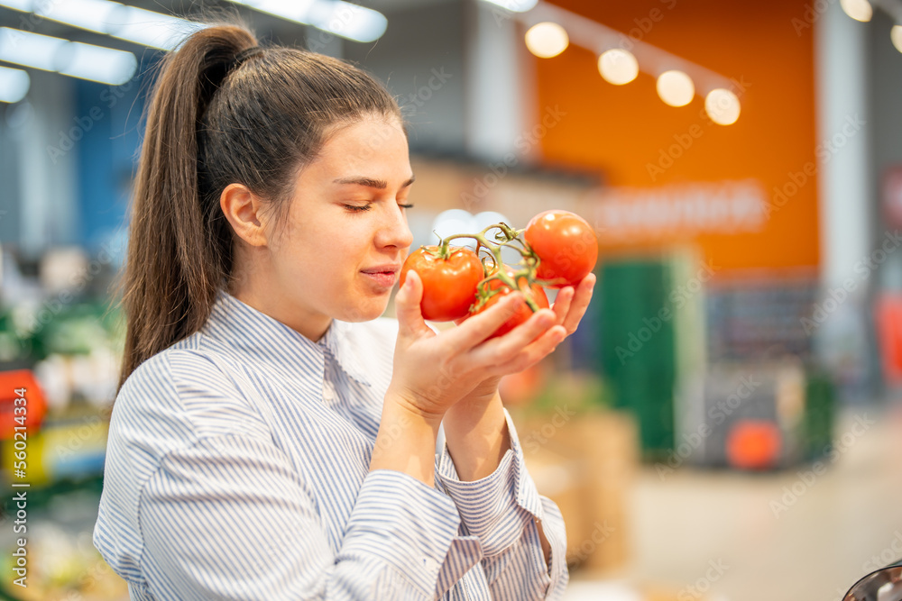 Beautiful young woman smelling fresh tomatoes with the eyes closed in vegetable section of grocery store. Woman choosing organic food during weekly shopping at the supermarket store.