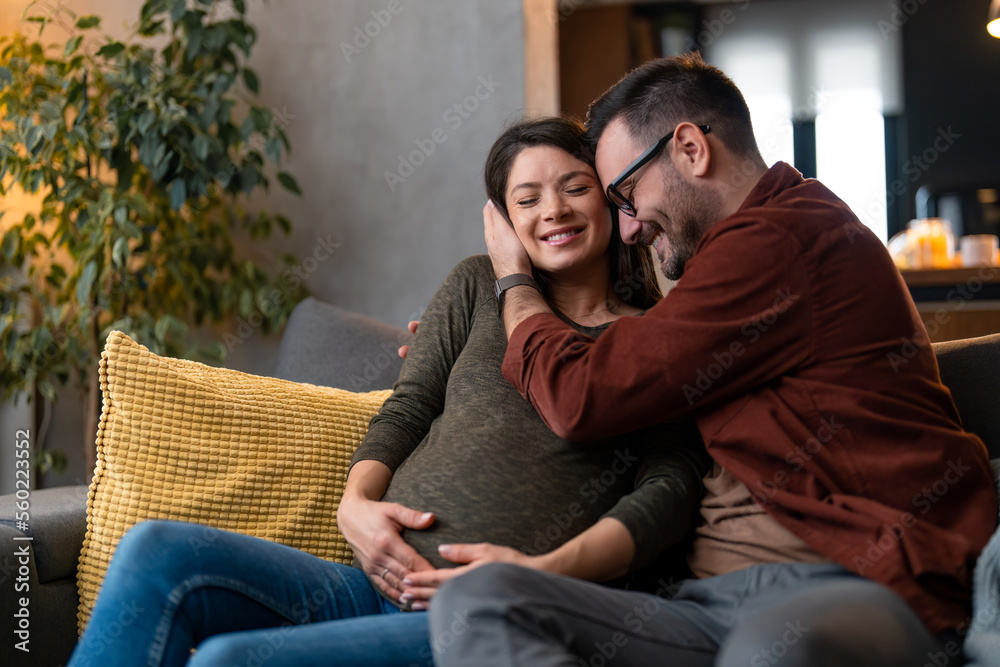 Supportive and caring husband gently hugging his pregnant wife at home while spending time together at home, sitting on a sofa, enjoying in peaceful moments. Supportive and caring husband gently hugging his pregnant wife at home while spending time together at home, sitting on a sofa, enjoying in peaceful moments.