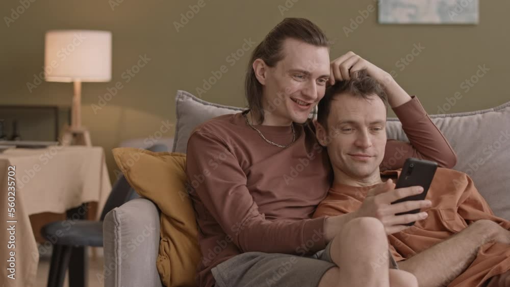 Happy peaceful gay couple spending time together at home relaxing on sofa in living room watching photos on smartphone Happy peaceful gay couple spending time together at home relaxing on sofa in living room watching photos on smartphone