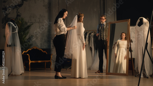 Cinematic shot of female tailor putting veil on woman in wedding dress in front of mirror. Bride on fitting wedding dress in designer atelier or wedding salon. Concept of fashion and couturier.