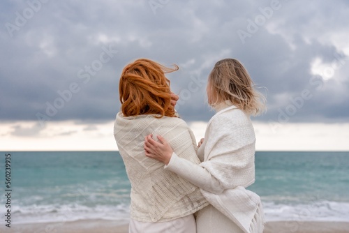 Women sea walk friendship spring. Two girlfriends, redhead and blonde, middle-aged walk along the sandy beach of the sea, dressed in white clothes. Against the backdrop of a cloudy sky and the winter 
