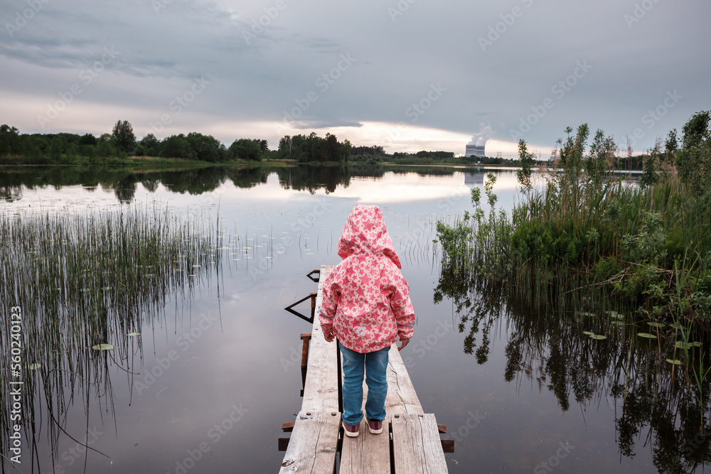A little girl in a pink jacket stands on a wooden pier, on the lake, at sunset. A little girl in a pink jacket stands on a wooden pier, on the lake, at sunset.