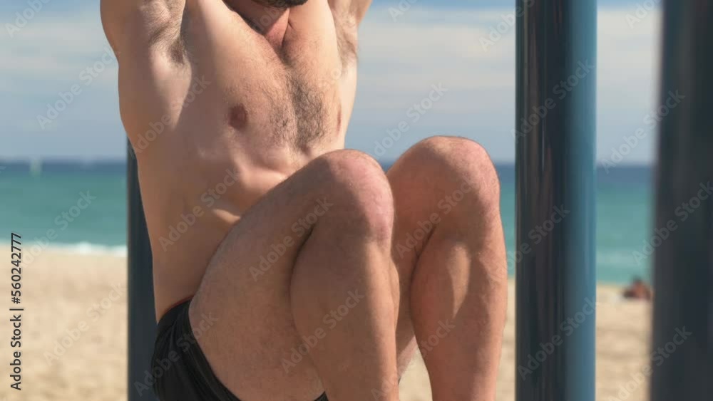 Shirtless young man doing sit-ups on a bar in a park at the beach.