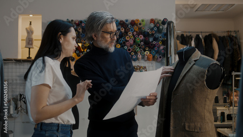 Male mature fashion designer discusses sketch drawing of future suit jacket with seamstress and shows tailoring process on mannequin. Atelier workshop on background. Fashion and hand craft concept.
