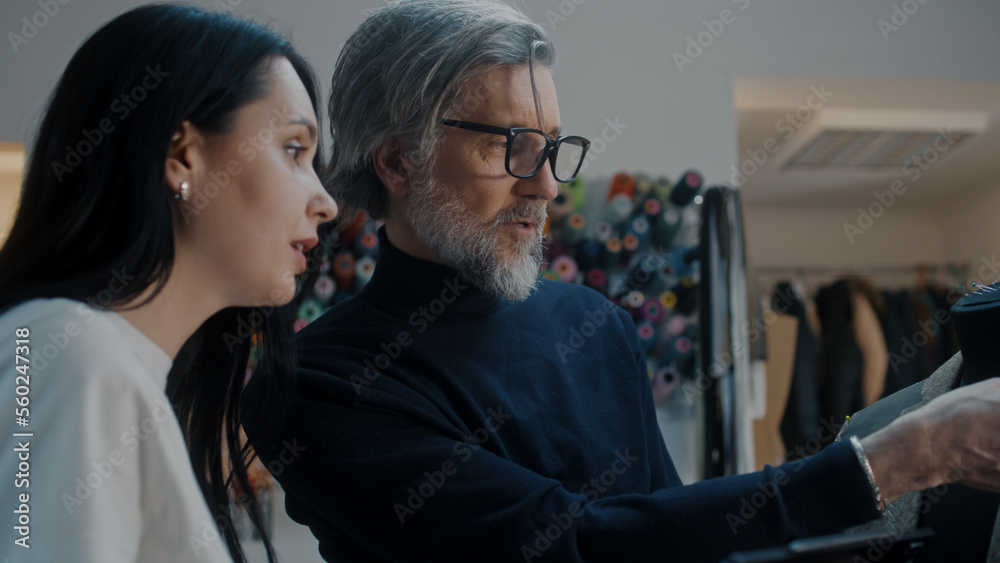 Male mature fashion designer with digital tablet talks with seamstress about future jacket and shows tailoring process on mannequin in atelier workshop. Fashion and hand craft concept. Close-up shot. Male mature fashion designer with digital tablet talks with seamstress about future jacket and shows tailoring process on mannequin in atelier workshop. Fashion and hand craft concept. Close-up shot.