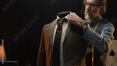 Male mature tailor works on elegant business or wedding suit. Mannequin with tailored shirt, tie and jacket in stylish luxury designer atelier or tailoring dim studio. Fashion and hand craft concept. photo