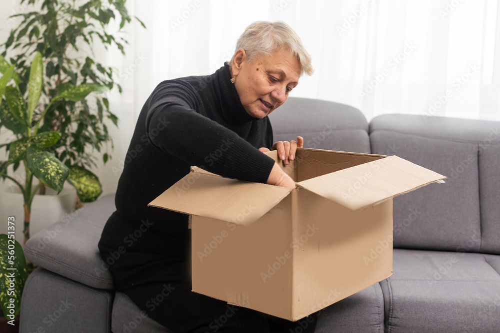 Beautiful senior woman is holding cardboard box sitting on sofa at home.