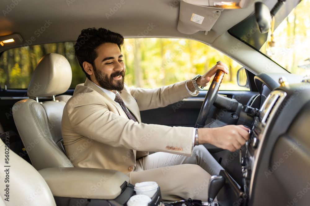 Happy arab young man businessman sitting inside luxury automobile, turning on music on dashboard before driving, handsome arabic man having car trip, side view, copy space Happy arab young man businessman sitting inside luxury automobile, turning on music on dashboard before driving, handsome arabic man having car trip, side view, copy space