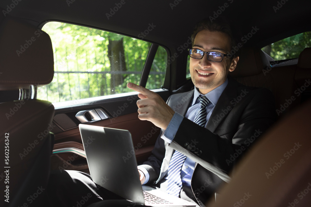Successful handsome businessman sitting in the passenger seat in his luxury car working on laptop and driving to a business meeting Successful handsome businessman sitting in the passenger seat in his luxury car working on laptop and driving to a business meeting