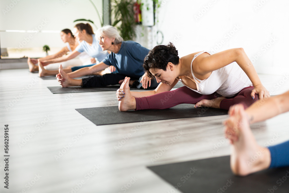 Concentrated young girl making stretching legs with group of active adult women sitting on yoga mat and doing exercise yoga together