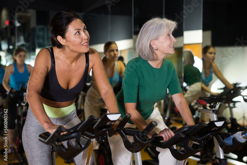 Portrait of young adult and mature women doing cardio workout at gym, training together on exercise bikes