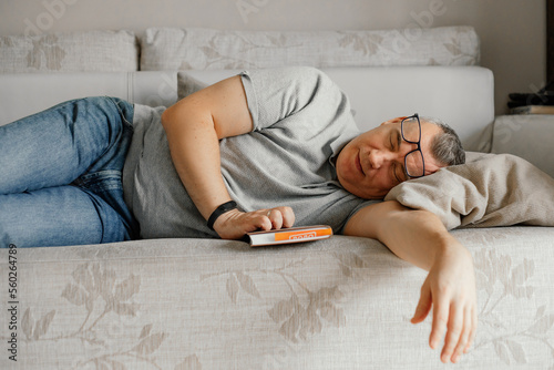 Aged man with glasses fall asleep reading book on comfortable couch in home room. Daytime sleep, napping.