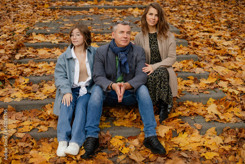 Happy family of three sit on stairs covered with fallen yellow leaves. Parents and teenage child walk in autumn park.