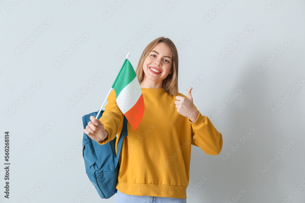 Young woman with flag of Ireland and backpack showing thumb-up on grey background