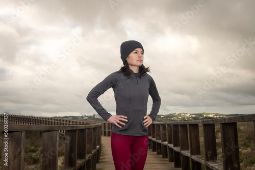 Full length shot of a woman standing with her hands on hips and looking away. Female athlete taking a break from running on a boardwak. © Rob Wilkinson