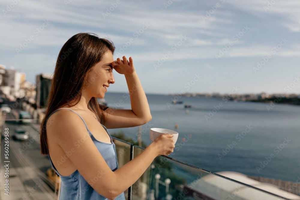 Close-up of beautiful brown-haired woman is standing on balcony and looking at sea with morning coffee on sunny day. Stylish cute lady in home clothes enjoying morning. People and free time concept