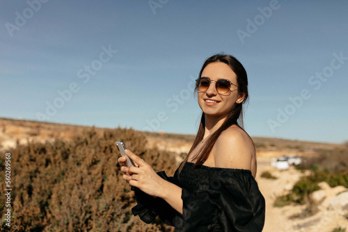 Charming adorable stylish woman with happy smile and dark hair wearing black dress with bare shoulders in sunglasses using smartphone and posing at camera while travelling in mountains 