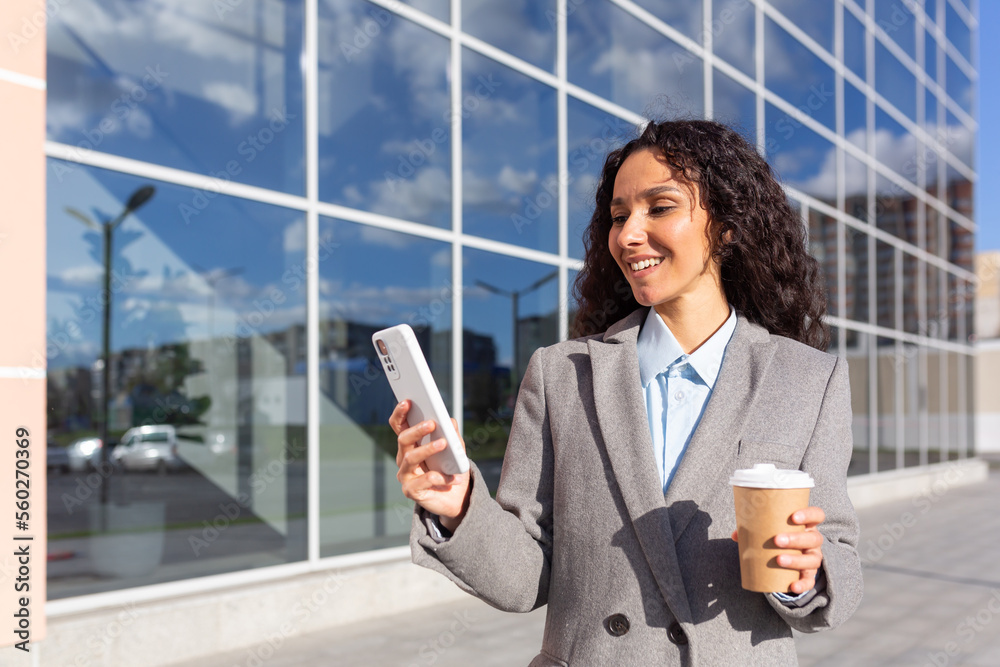 Beautiful and successful hispanic business woman using smartphone from outside modern office building, woman with a cup of hot drink walking down the street. Beautiful and successful hispanic business woman using smartphone from outside modern office building, woman with a cup of hot drink walking down the street.