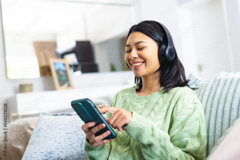 Image of happy biracial woman in headphones using smartphone sitting on sofa at home, copy space Image of happy biracial woman in headphones using smartphone sitting on sofa at home, copy space