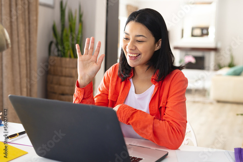 Image of happy biracial woman sitting at desk waving during video call on laptop at home, copy space