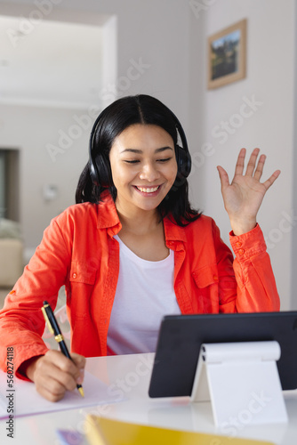 Vertical of happy biracial woman in headphones waving, making tablet video call at home, copy space