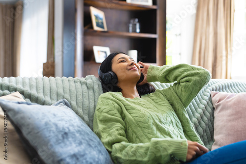 Image of happy biracial woman in headphones, relaxing on sofa with eyes closed at home, copy space