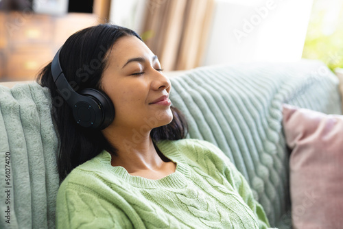 Image of happy biracial woman in headphones relaxing on sofa with eyes closed at home, copy space