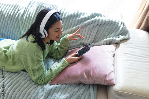 Image of happy biracial woman in headphones using smartphone lying on sofa at home, copy space