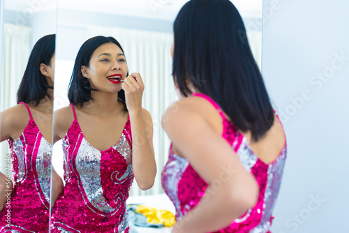 Happy biracial woman in party dress looking in mirror, putting on lipstick, copy space
