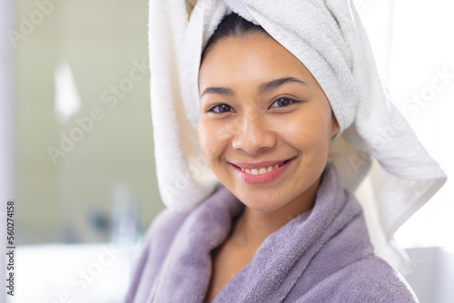 Portrait of happy biracial woman wearing bathrobe and towel smiling in bathroom, with copy space