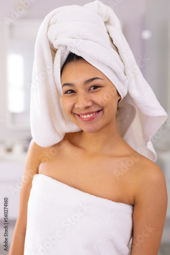Vertical portrait of happy biracial woman wearing towel smiling in bathroom, with copy space