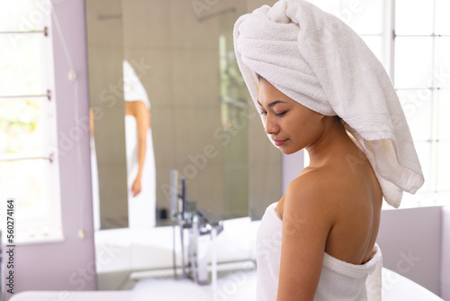 Happy biracial woman wearing towel running bath in bathroom, with copy space
