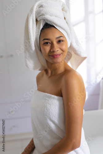 Vertical portrait of happy biracial woman wearing towel, smiling in bathroom, with copy space