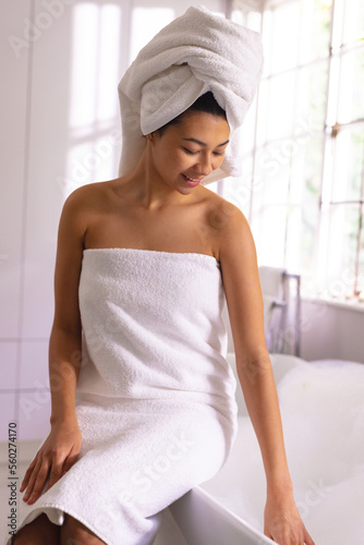 Vertical of happy biracial woman wearing towel sitting on the edge of bath smiling, with copy space