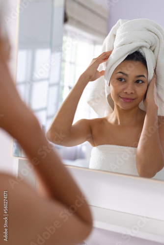 Vertical of happy biracial woman adjusting towel on head smiling in bathroom mirror, with copy space