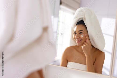 Happy biracial woman wearing towel smiling in bathroom mirror moisturising face, with copy space
