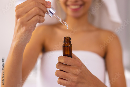 Smiling biracial woman wearing towel in bathroom, holding essential oil dropper bottle, copy space