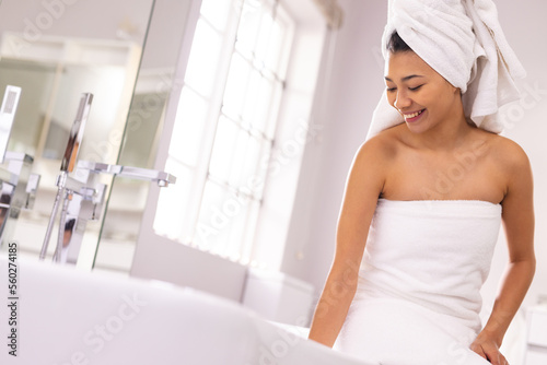 Smiling biracial woman wearing towel in bathroom, preparing bath, with copy space