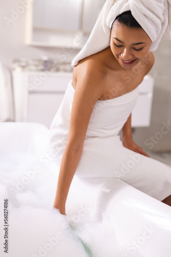 Vertical of smiling biracial woman wearing towel in bathroom, preparing foam bath, with copy space