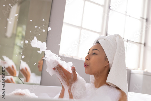 Image of biracial woman with towel on head blowing foam in bathtub in bubble bath in bathroom photo