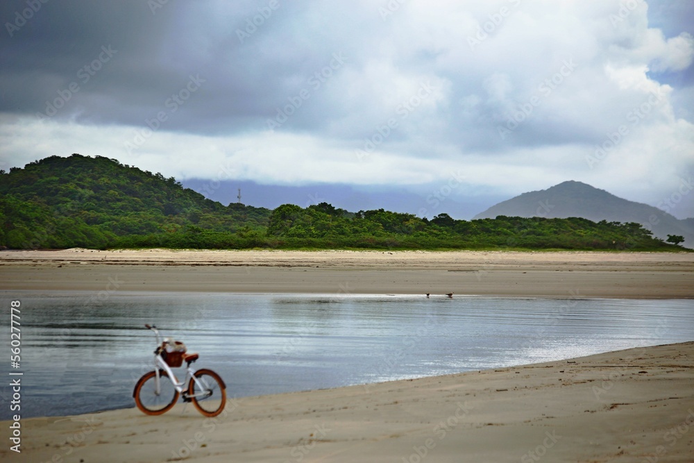 Obraz premium Bike standing on the sand of river side,, River with calm waters and green forest on the background. Itaguare beach, Bertioga, Brazil