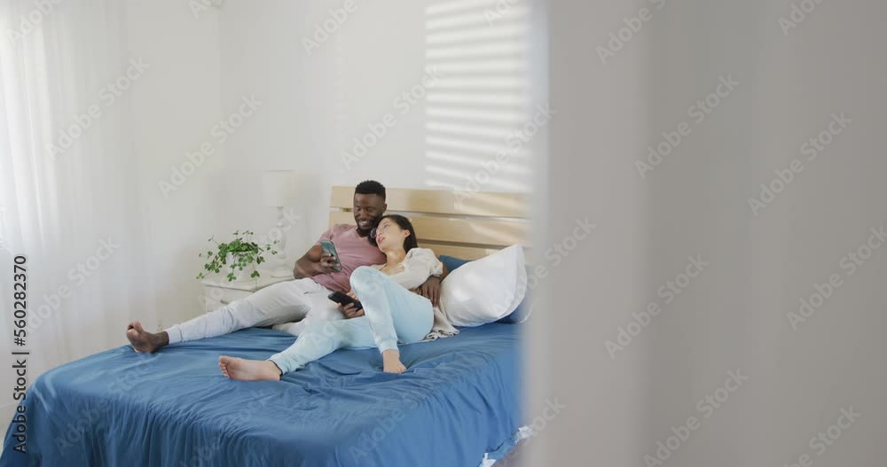 Happy diverse couple using smartphone and lying in bedroom Happy diverse couple using smartphone and lying in bedroom