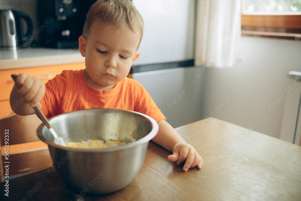the child cooks in the kitchen, stirring the dough in a metal bowl the child cooks in the kitchen, stirring the dough in a metal bowl