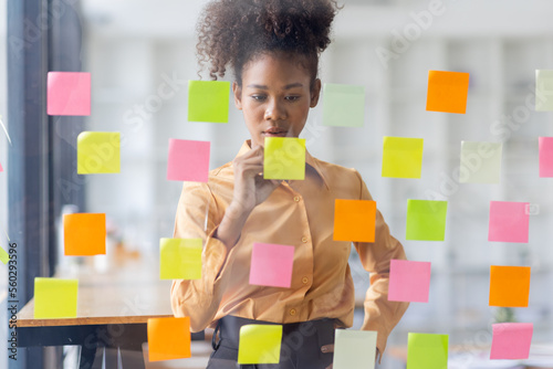 Young Serious african american Creative team use post it notes to share idea sticky note on glass wall. Asian business people design planning and Brainstorming thinking sticky History notes concept.