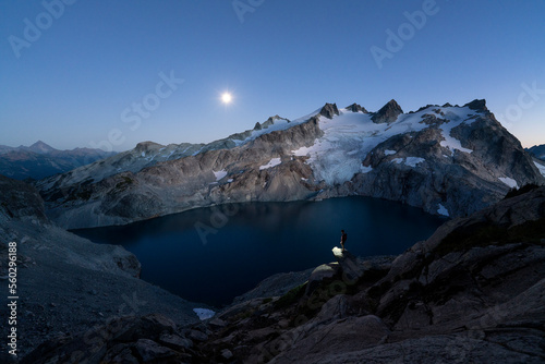 Man standing on ledge over alpine lake with headlamp on and the moon in the distance