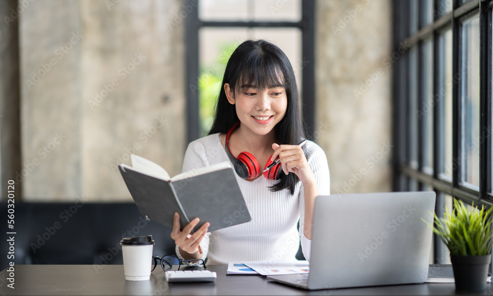 Fototapeta premium Happy Asian girl student wear wireless headphone study online with the teacher, happy young woman learn language listen lecture watch webinar write notes look at laptop sit in cafe, distance education