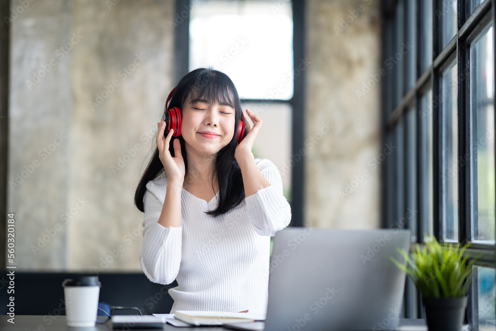 © crizzystudio - Happy Asian woman relaxing and listening to music in office with computer laptop and coffee cup. People and lifestyles concept. Freelance and outdoors workplace outdoors theme.