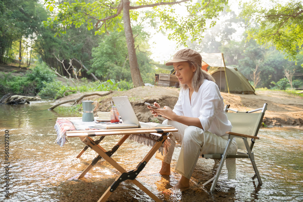 Asian woman travel and camping alone reading a book and using laptop while relaxing on the deck chairs in the river at natural park in Thailand. Recreation and journey outdoor activity lifestyle. Asian woman travel and camping alone reading a book and using laptop while relaxing on the deck chairs in the river at natural park in Thailand. Recreation and journey outdoor activity lifestyle.